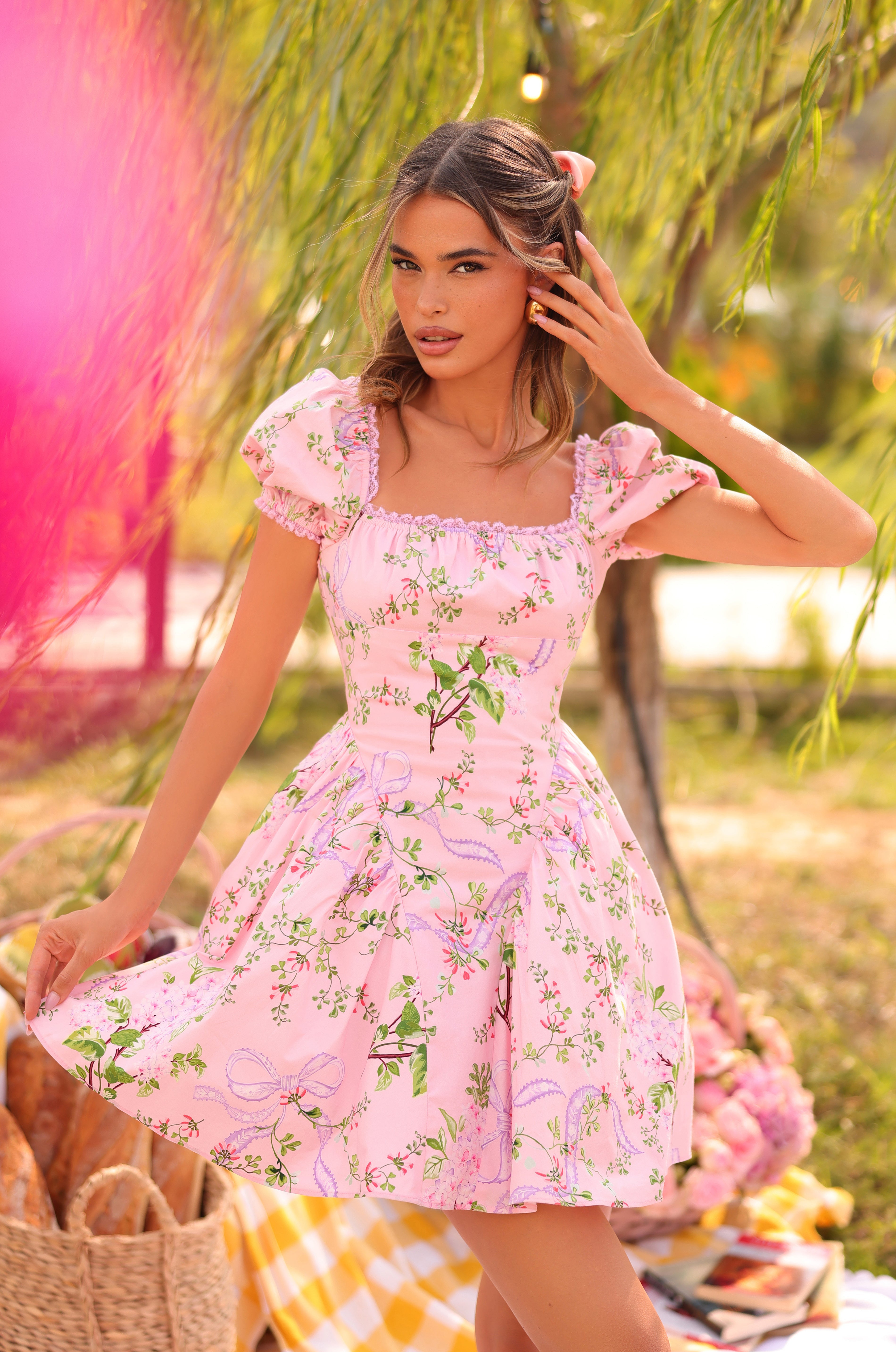 Outdoors near a picnic with bread and flowers, a woman wearing the Blush Bloom Floral Short Casual Dress: Rosabella holds one side of her blush bloom skirt amid trees and greenery.